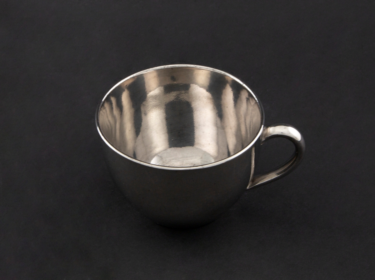 Kopp i keramikk, glasert med sølvfarget luster. Tilnærmet sylinderformet korpus med påsatt hank. Mangler skål.
