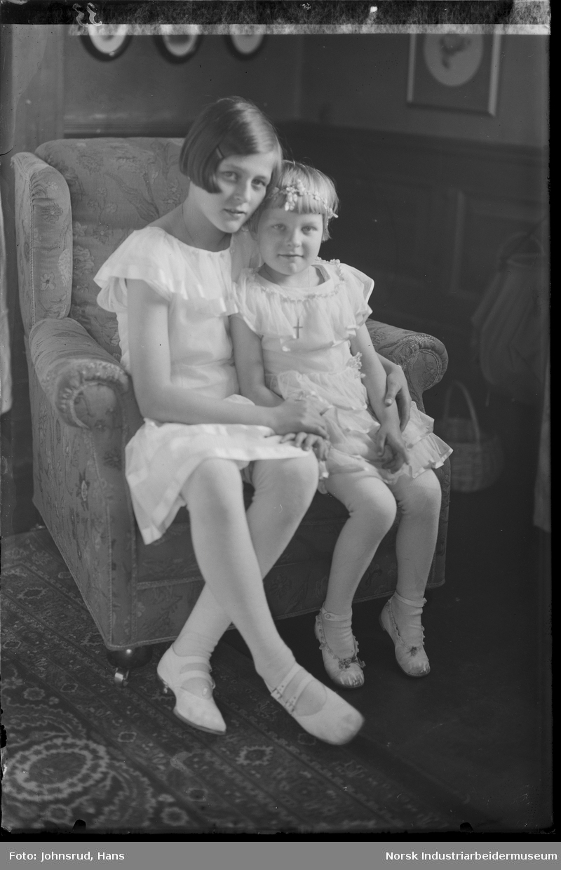Gruppeportrett. To barn sittende på stol i stue. Begge kledd i kjoler.
Portrett bestilt av Direktør Hansen.
