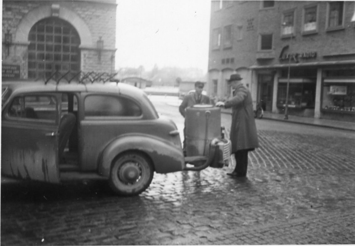 Två män står vid ett gengasaggregat som sitter i bakänden på en personbil. Det är lite blöt på marken efter ett regn. I bakgrunden en större tegelbyggnad med väggskylt: "Luxor Radio". De befinner sig på Hoppets torg i Jönköping.