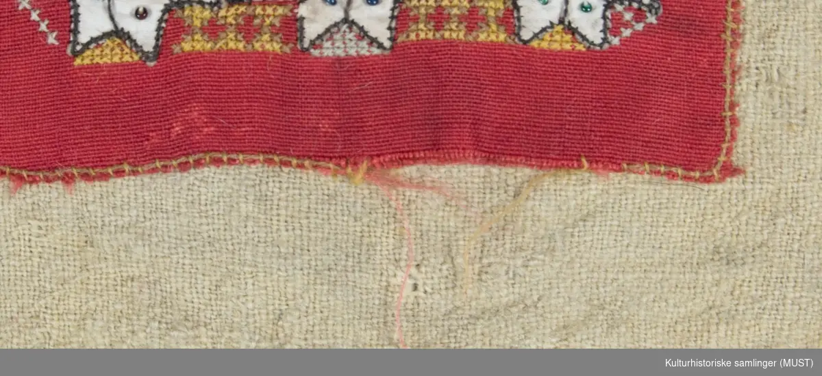 Brystduk, rød med utsaum i sort og hvitt og andre farger. 8-bladsroser og annen pynt. Øverst gullbånd.
