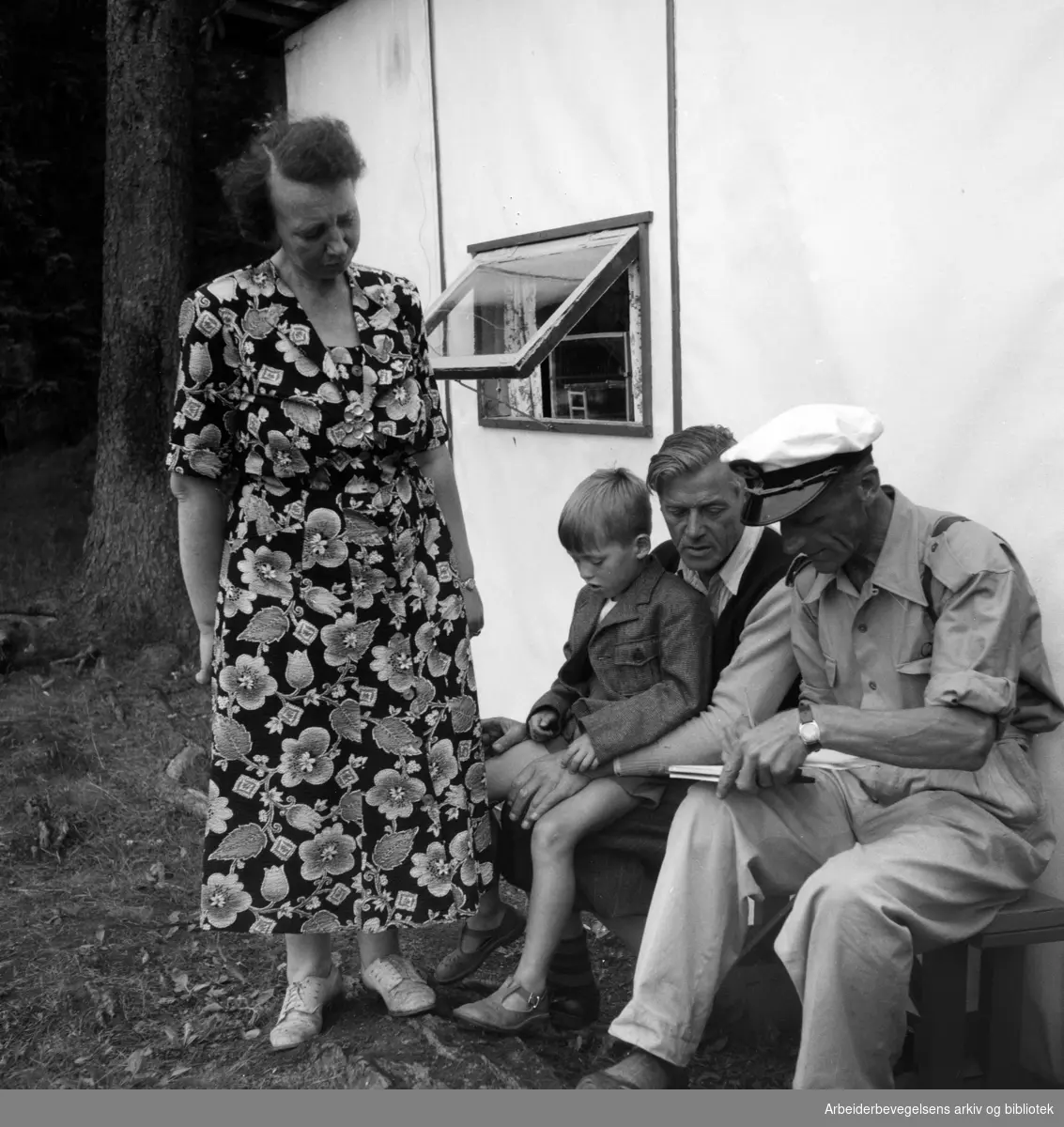 Georg Larsen og familie tilbringer sommerferien på Nordre Langåra i en 9 kvadratmeter stor hytte laget av papp. Vaktmann Rolf E. Nilsen er på besøk for å ta opp "manntal". Juli 1954