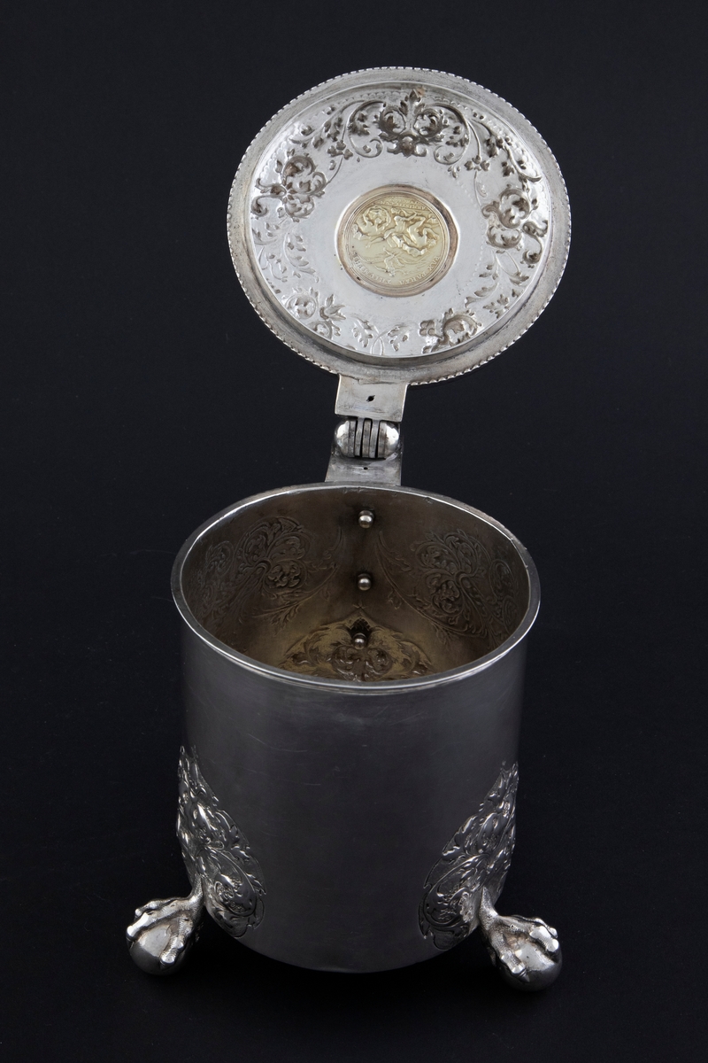 Drikkekanne i sølv, delvis forgylt, med sylindrisk korpus, hank og hengslet lokk. Føttene er tre kuler med klo. Akantusdekor på korpus og lokk. Lokkåpner er formet som sittende løve med kule. Lokket har innfelt medaljong med bilde av kong Leopold I av Det tysk-romerske rike (1640-1705), omkranset av akantusranker. På innsiden av lokket er medaljongen dekorert med motiv som viser titanenes fall.