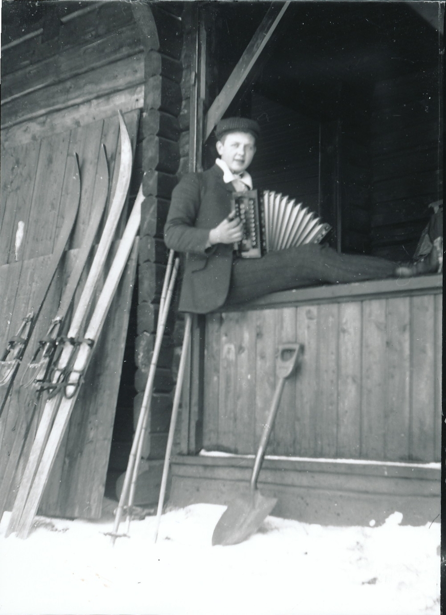 Harald Rydgren sitter og spiller trekkspill på verandagelenderet utenfor Mosjøhytta i Nordmarka. Ski, staver og spade oppstilt mot hytteveggen.