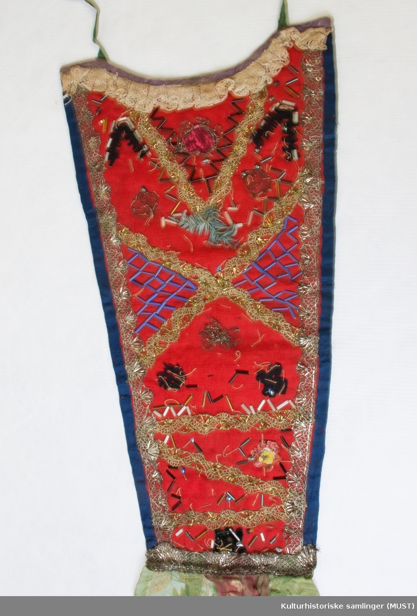 Dåpssmekke av rødt tøy kantet med blå silke, gull- og sølvborder og perlepynt. Grønne og røde pyntebånd. For av blåmønstret tøy.
