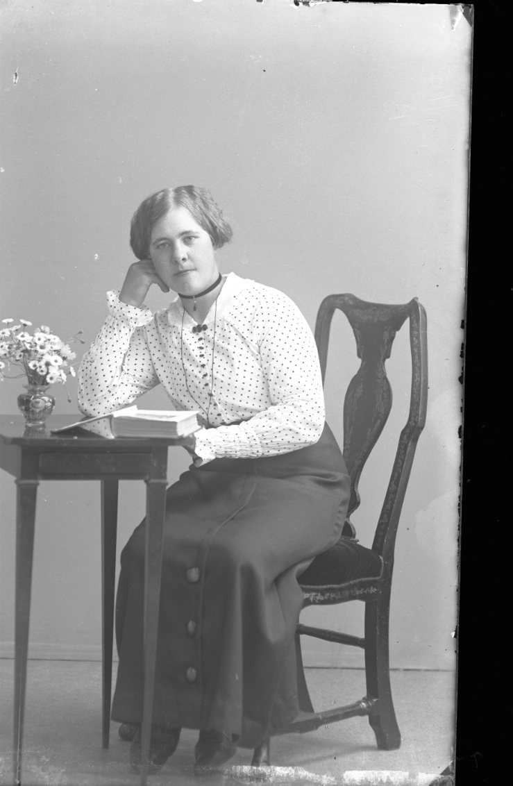 En ung kvinna i prickig blus och mörk klänning sitter vid ett bord med en uppslagen bok. På bordet står också en vas med blommor.