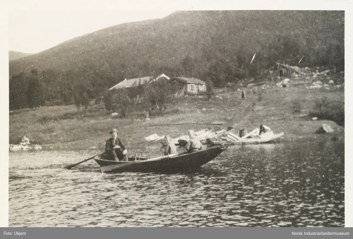 Fra Liset. En mann med hatt og to gutter i en pråm på Møsvatn. Personene ror båten ved siden av land. På land ligger flere båter langs vannkanten, og et småbruk