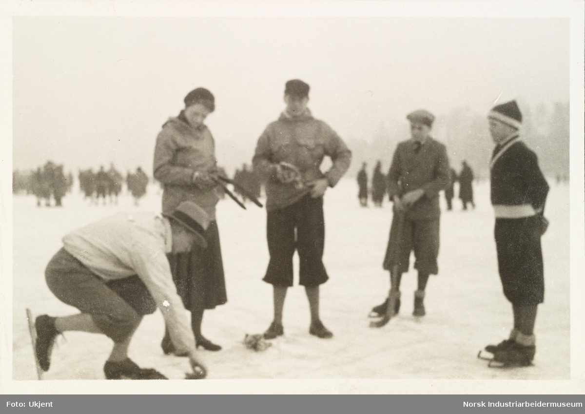 Fra Holtekilen, januar 1932. En  mann med skøyter på bena lener seg ned på isen. Fire andre, en kvinne og tre menn ser på han. Flere av personene har også på skøyter. I bakgrunn flere mennesker på isen.