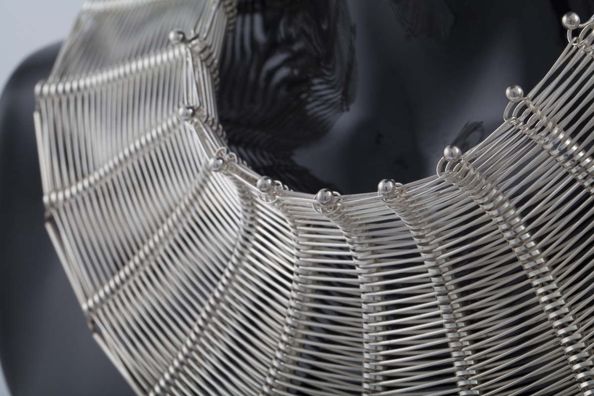 Klave i sølv, utført i en teknikk der kunstneren har latt seg inspirere av tekstilkunst. Smykket vokser tilsynelatende ut fra halsen, hvor det oppstår et spill mellom radiære spiralformer og de horisontale sølvelementene. Smykket lukkes i nakken med sølvknapper og bølgeformede hekter.