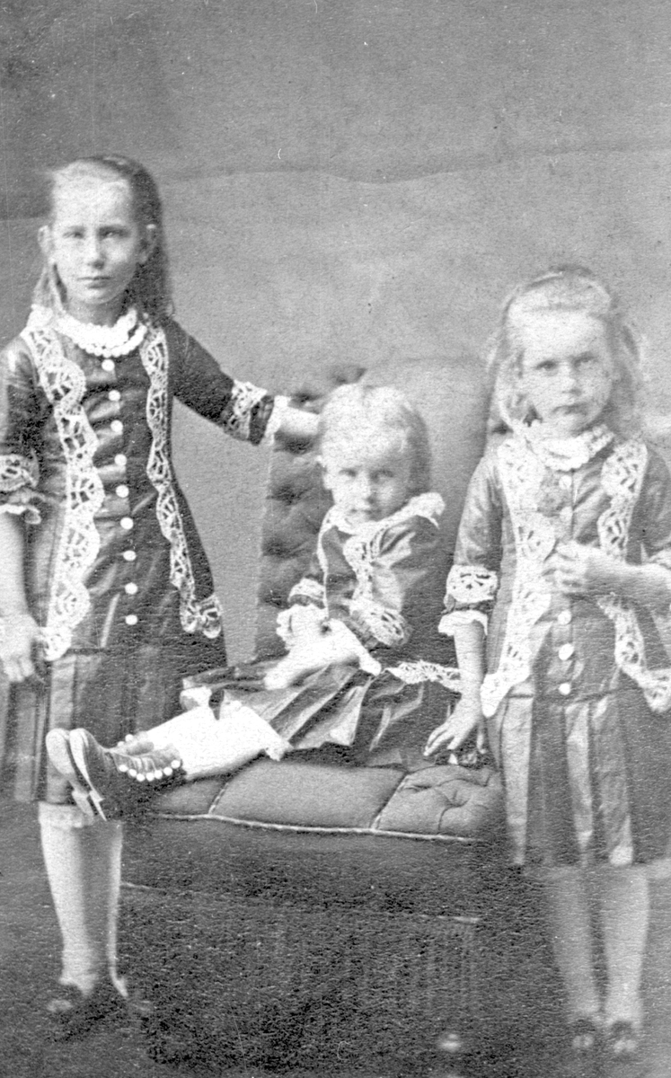 Portrett av tre jenter. Alle har like kjoler, pyntet med blonder og med foldeskjørt. Hvite knestrømper.
