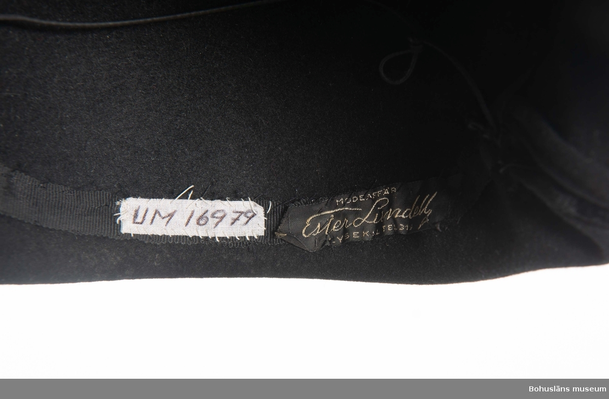 Svart formpressad hatt. Prydd med svart sammeband. 
Isydd band med text: "Modeaffär Ester Lundell Lysekil tel 319."