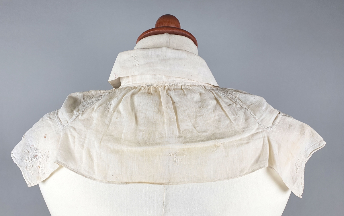 Skjortebryst av transparent bomull med krage. Broderte blomster i hvit tråd av plattsøm både på kragen, brystet og på ryggen. Stoffet dekker hele brystet og kun øverste del av ryggen.