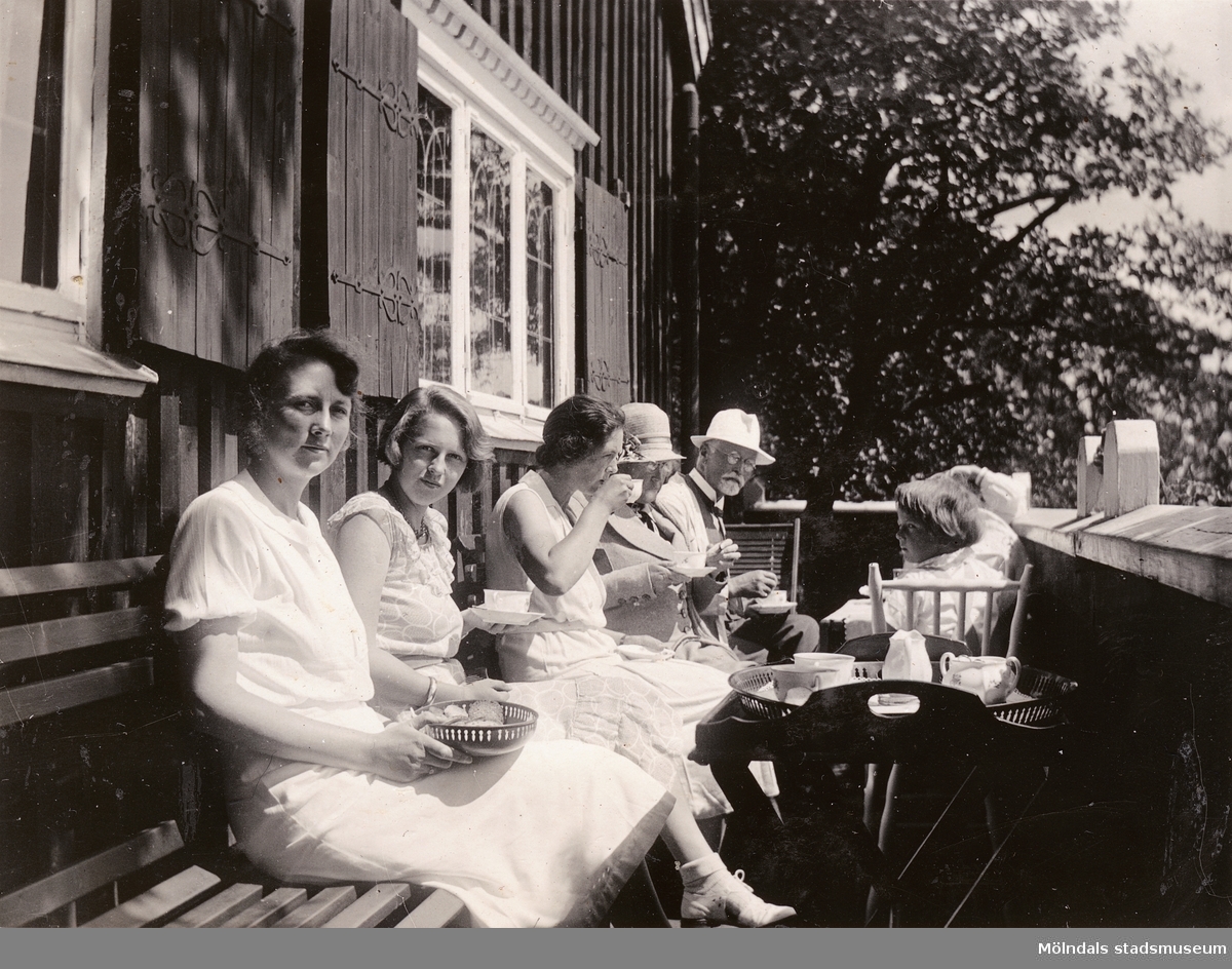 En samling personer dricker kaffe på verandan som vetter mot Landvettersjön. Okänt årtal.

Fotografi taget vid sommarbostaden Strandlyckan, Slamby 1:4 i Landvetter socken. Huset övertogs senare av Stiftelsen Enandergården, Härlanda församling, på 1950-talet.