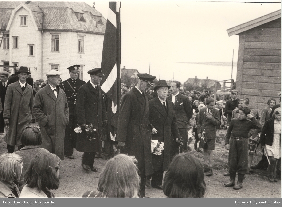 Kong Haakon VII på besøk i Vadsø i 1946. Kong Haakon går først i et tog. Til høyre side går fylkesmann Hans Julius Gabrielsen. Mange mennesker har kommet til stede for å se på kongen. Vadsø sykehus (kysthospitalet) i bakgrunnen. Se også bildene 264-267.