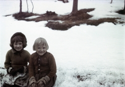 To unge jenter sitter på en snøkant i et snødekt landskap.
