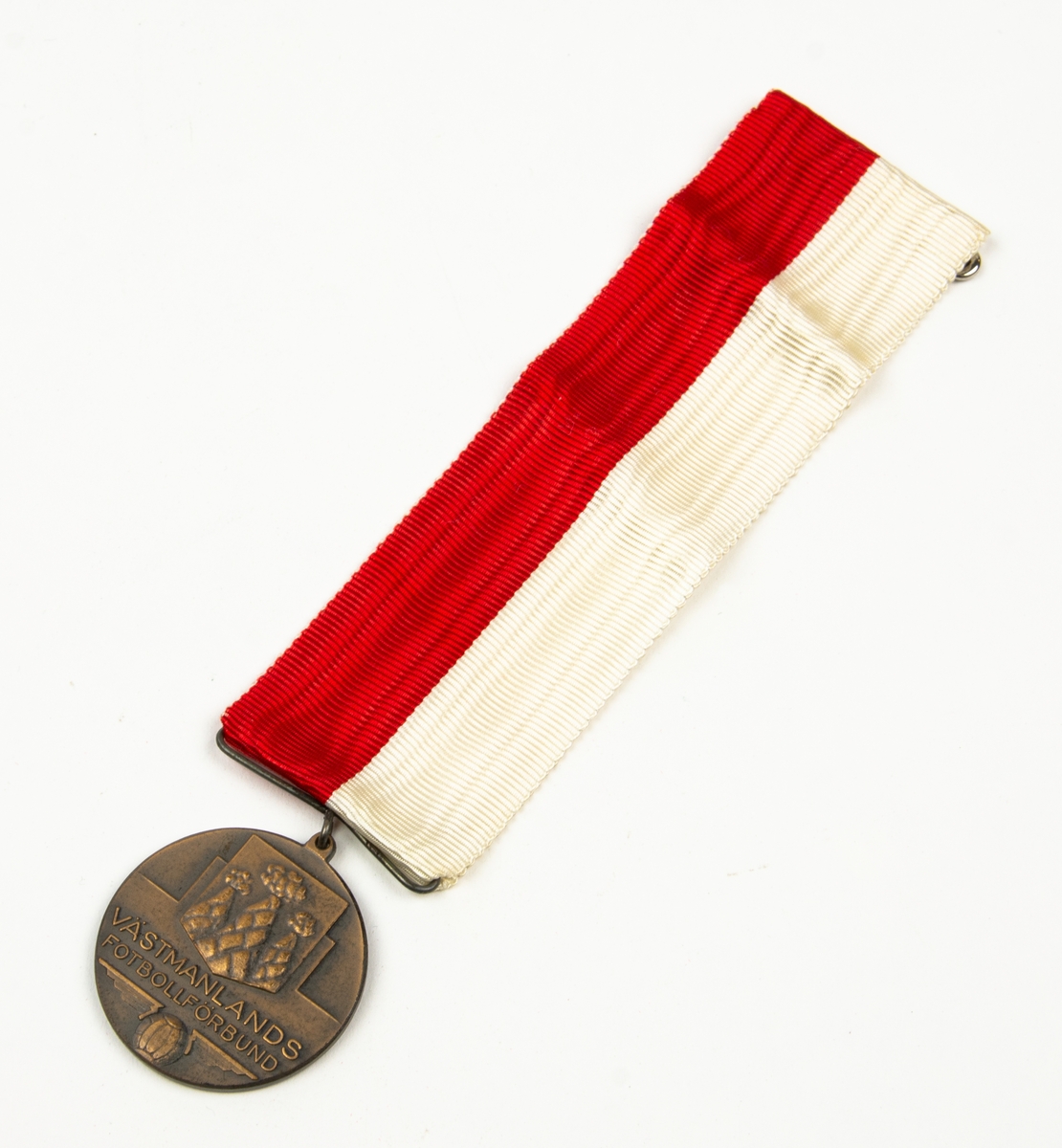Medalj med band i vitt och rött. På medaljens ena sida finns en sköld med tre milor. Under skölden finns texten: VÄSTMANLANDS FOTBOLLSFÖRBUND På medaljens andra sida finns texten: DIV. 7 V:ÅS GRUPPEN 1955.