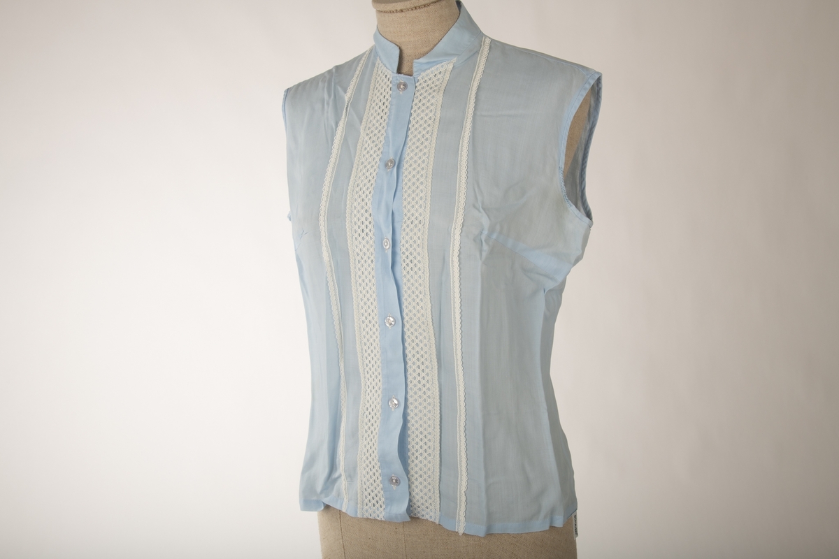 En lyseblå ermeløs bluse, med hvite kniplede bånd sydd på i front. Krage uten brett og knapper og knappehull foran.