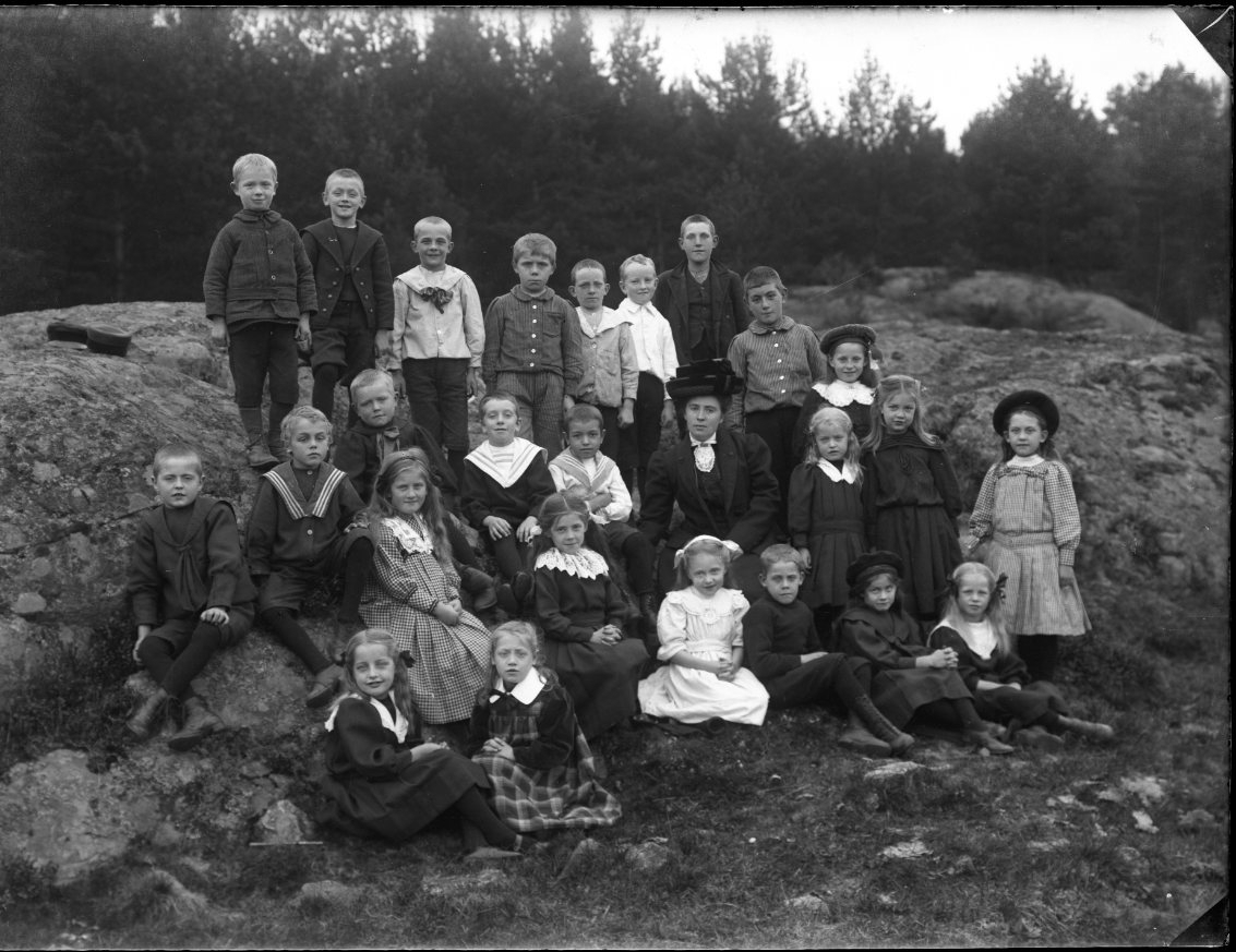 En skolklass med flickor och pojkar fotograferade vid en bergknalle. Lärarinnan heter sannolikt Bengtsson.