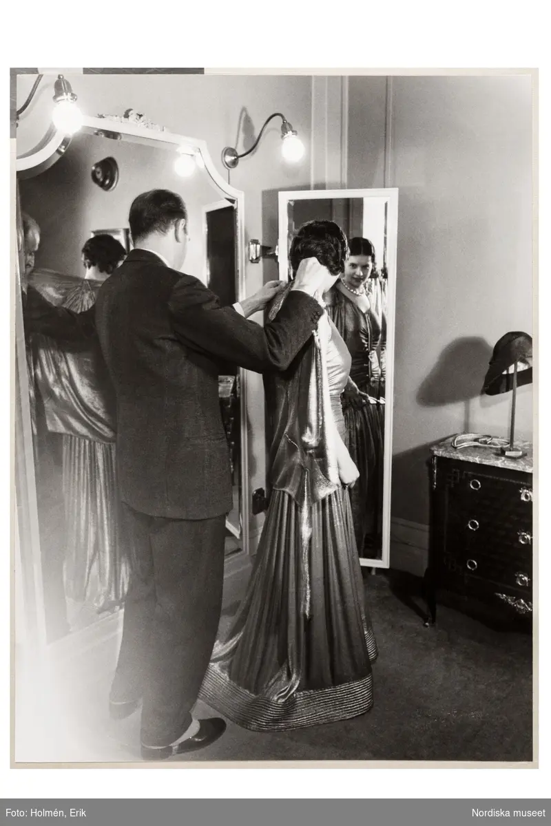 Mannekäng visar mode vid speglar i provrummet. En man, Pelle Lundgren (1896-1974) hjälper henne på med jackan. Festklänning med rynkat liv, hellång svagt utställd kjol med dekor nedtill. NK:s Franska damskrädderi.