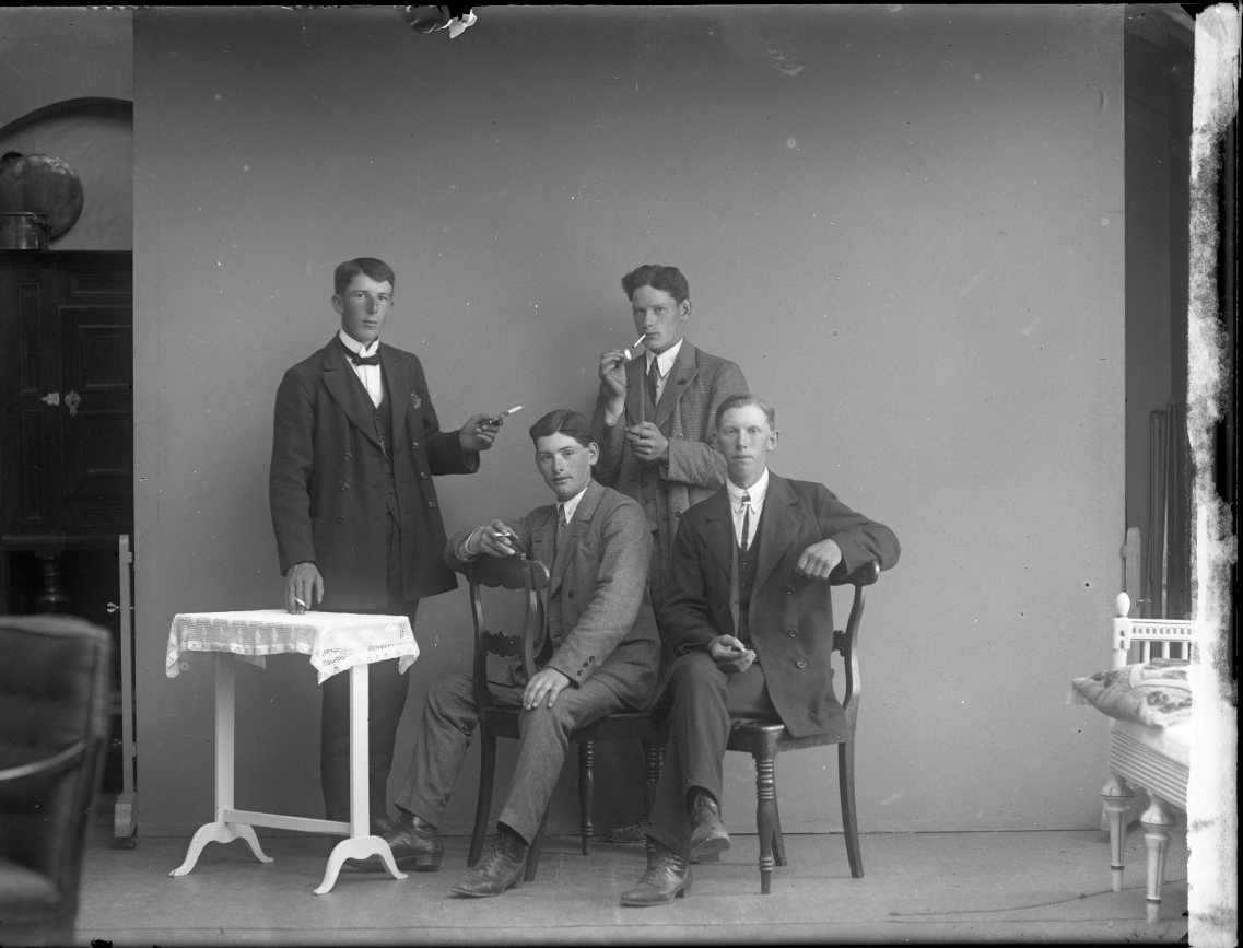 Fyra unga män med cigaretter i händerna. Några står andra sitter, bland annat bak-och-fram på en stol. Möjligen mönstrinsbild. En av männen är Mandor Johansson från Västanå, Gränna socken.