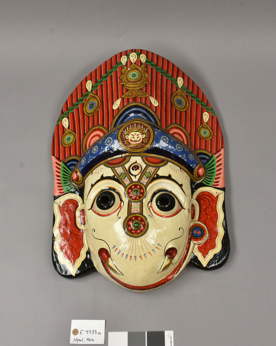 E 9737 a Fra protokoll: Maske av leire, bomull, strie, rispapir, bomullstau og maling. Støpt på form (i leire), tørket, forsterket,  kalket og malt.#Figurens navn: /Ganay dyah/ (=Ganesh). Hinduistisk gud. En i ett sett på 13 masker. Brukes i religiøst drama kalt /Nawa Durga Pyájhan/ (= Ni Durga Dansen). Tilbedelses objekt. Masken er kopi av original som ikke kan kjøpes fordi de brennes i et esoterisk rituale på slutten av hver danse sesong. (8 mndr. dansing, 4 mndr. pause) Brukes av mannlige dansere fra blomstersamlerkasten; /Gátha/.#Produsent: Purna Chitrakar m/fam. Malerkastefam.
