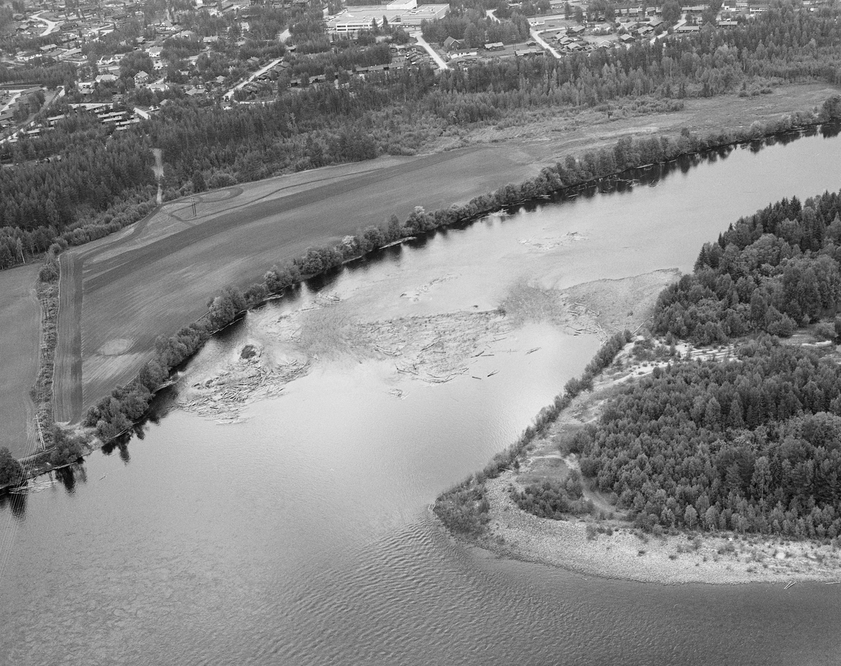 Flyfotografi tatt over Glomma, ved nordenden av Mariholmen, der vi ser hvordan ien del tømmerstokker hadde satt seg fast i elveløpet på østsida av holmen. På østsida av elva ser vi åkerarealene på Strandmoen og villabebyggelsen på Strandstykket.