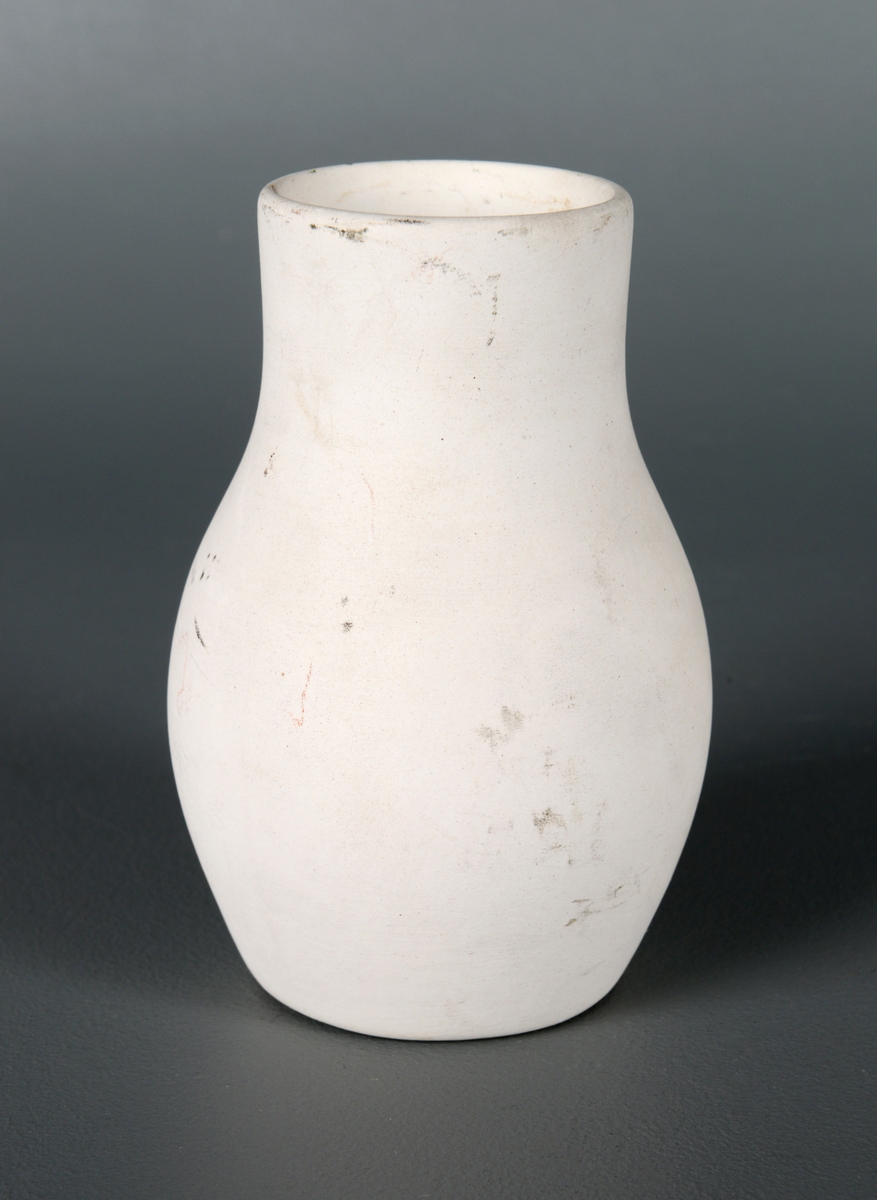 En liten blomstervase av hvit, uglasert fajanse. Vasen har sirkulær bunn of rund form. Øverst smalner den inn til en ganske bred og rett hals. Vasen har ingen dekor, men det er en inskripsjon hvor det står "56" på undersiden.