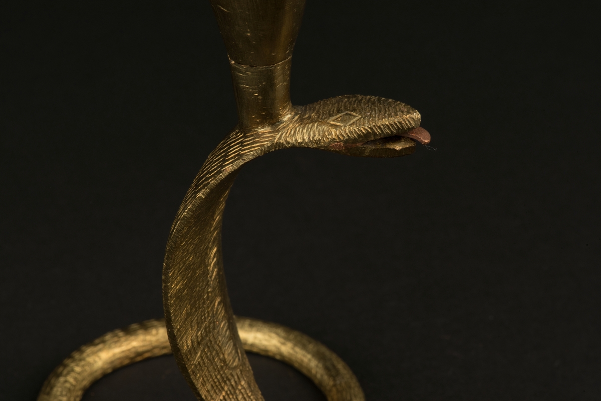 Ljusstake av mässing i form av kobraorm.
Ormens huvud är rest och svansen fungerar som stöd och fot. 
Ljushållaren är fäst på ormens huvud med en skruv.
