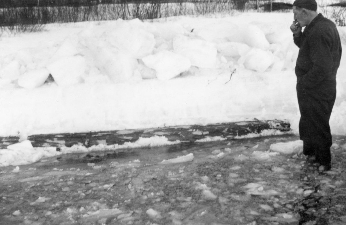 Tømmer som har vært nedfrosset i isen på Glomma ved Støeng hogges løs.  Fotografiet viser en barket furustokk av finerkvalitet som er hogd løs, og som ligger og flyter i isvann.  En mann med kjeledress, gummistøvler, topplue og sigarett i munnen betrakter det hele fra en posisjon til høyre i bildeflata.  I bakgrunnen snødekt is med en opphopning av digre isflak.  Bakenfor dette igjen en elvebredd med krattskog. 