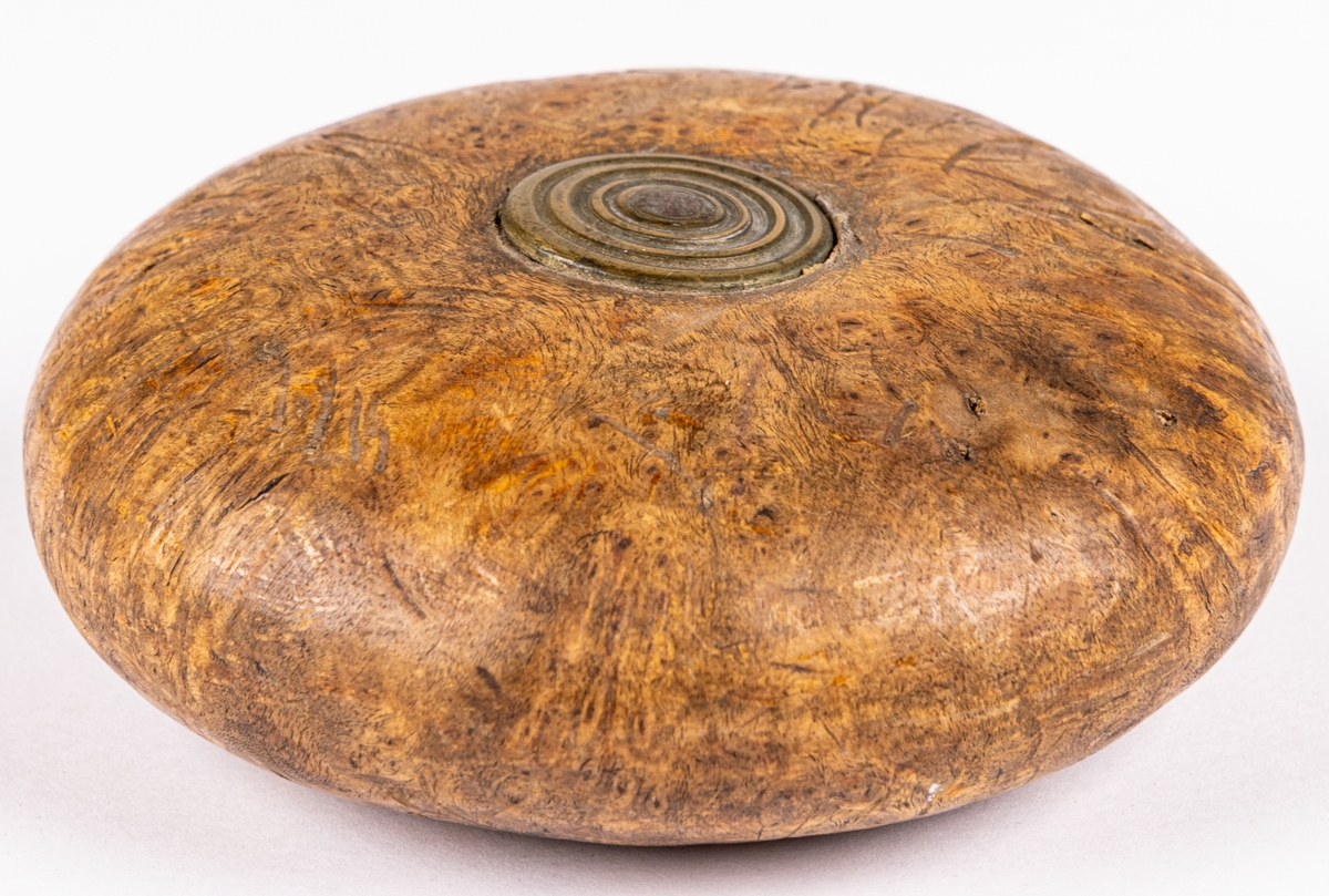 Snushorn, av masur, svarvat, rundat, platt, med ett runt mässingsbeslag på mitten av båda plattsidorna. I ena kanten hål för träplugg.