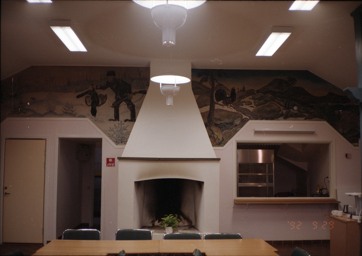 Interiör med dörr, eldstad och öppning in till ett kök i byggnaden "Tjädern". Väggmålning med motivet av en jägare med skjuten tjäder i handen, och stenigt landskap med tjäder i ett träd.