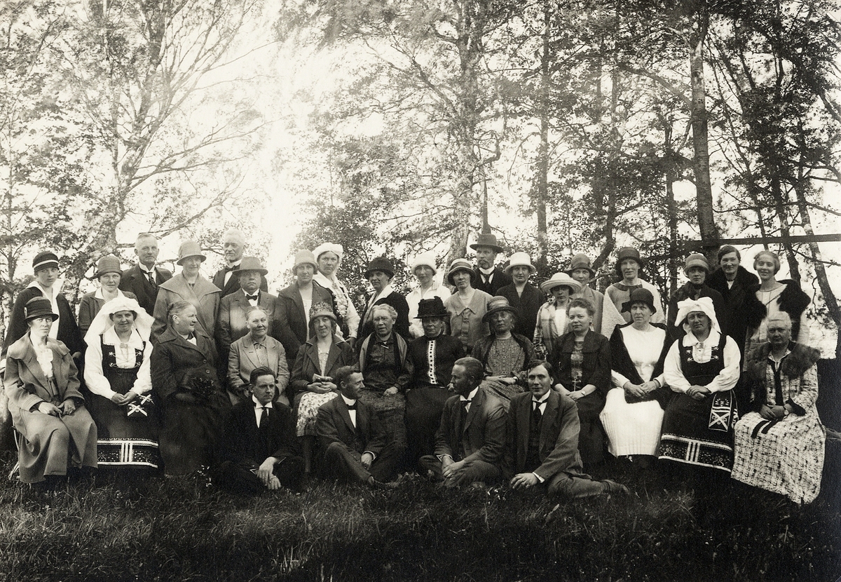 Enligt text: "Lärarkåren (kollegiet) vid Växjö folkskola på utflykt, 1925". 
Ett antal herrar och damer har slagit sig ner i en lövskogsslänt. Tre av kvinnorna är klädda i Värendsdräkt.