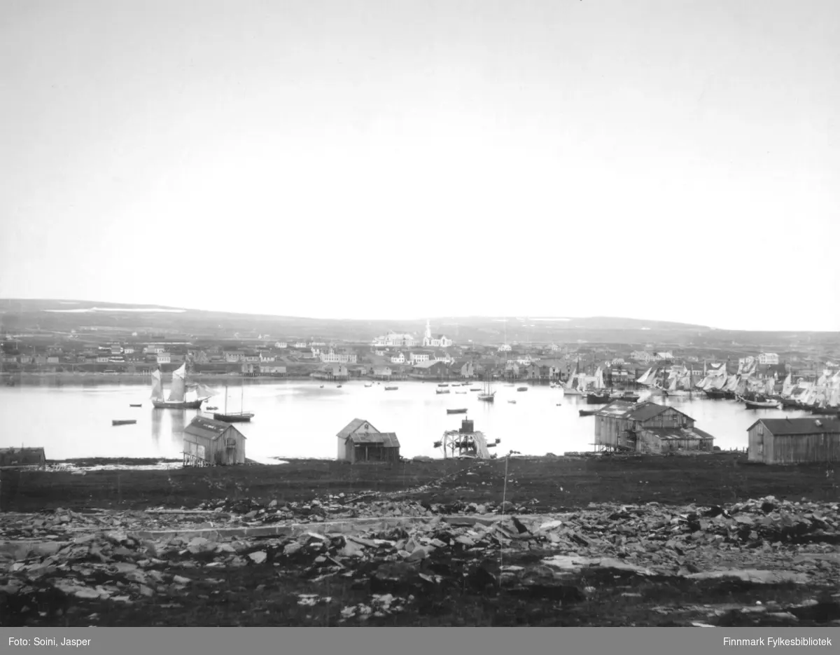 Et oversiktsbilde over Vadsø tatt fra Øya, montert på passepartoutkartong. På havna ser vi mange store seilskuter/pomorskuter, en dampbåt og mange nordlandsbåter.  Vi ser bl.a. Amtmannsgården og Vadsø kirke i bakgrunnen.