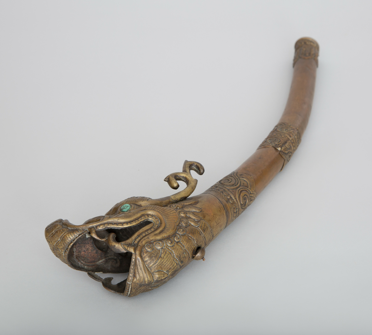 Blåseinstrument av kobber, med munnstykke, tverrbånd og klokkestykke av ornamentert messing. Klokkestykket har form av et dragehode med oppsperret gap. Øyer av turkis edelsten.