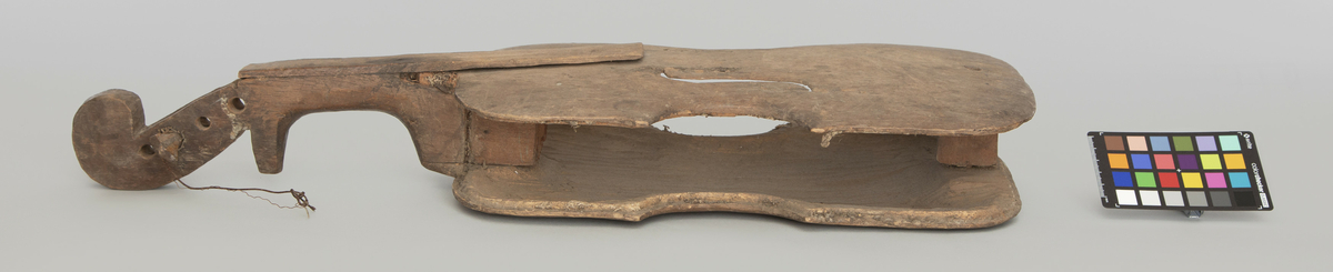 Fragment. 4-strenget. Laget av ubehandlet tre, skåret med kniv. Meget primitivt. Form omtrent som en vanlig fiolin, men svært lite hvlevet bunn og lokk.
