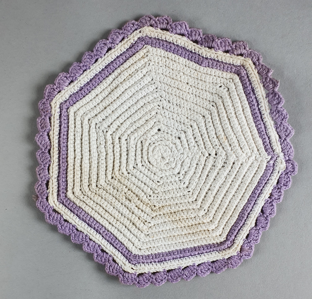 Femkantet - nesten rund - heklet pyntebrikke av bomull med lilla dekorkant.