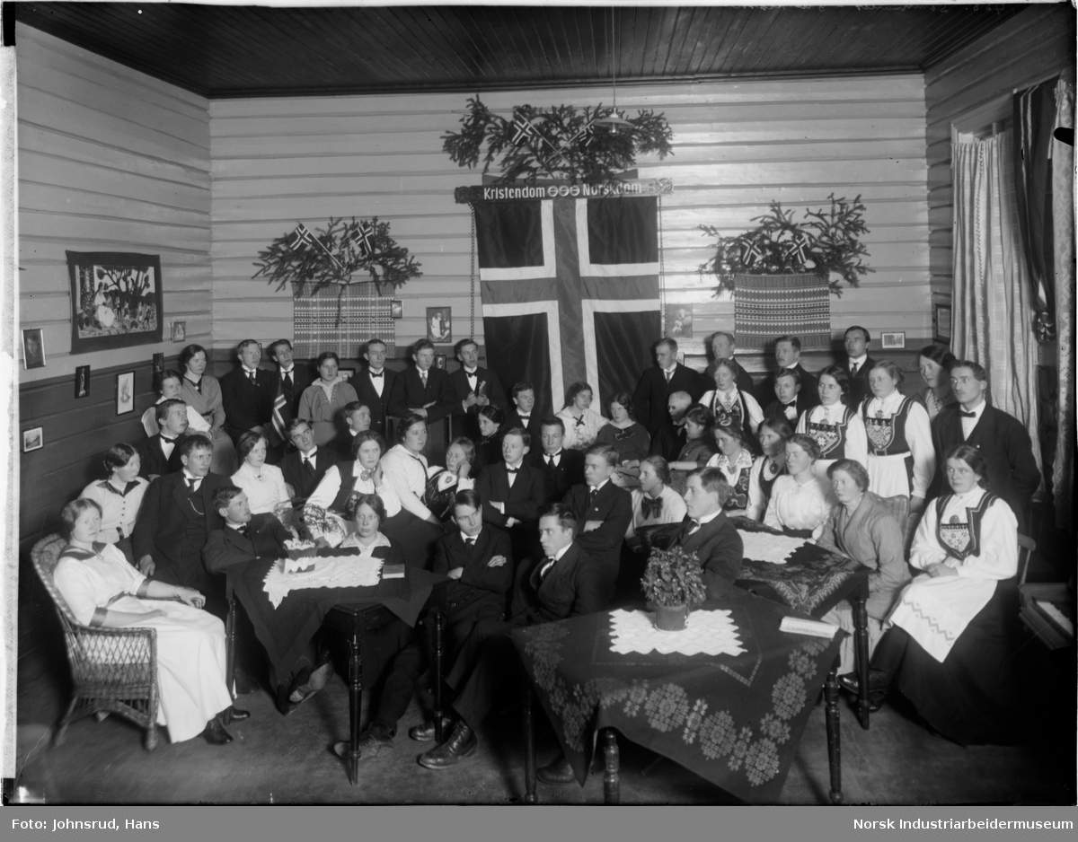 Gruppebilde av tredje klasse 1915-1916 ved seminaret forografert i en stue. På veggen bak er det et stort norsk flagg.