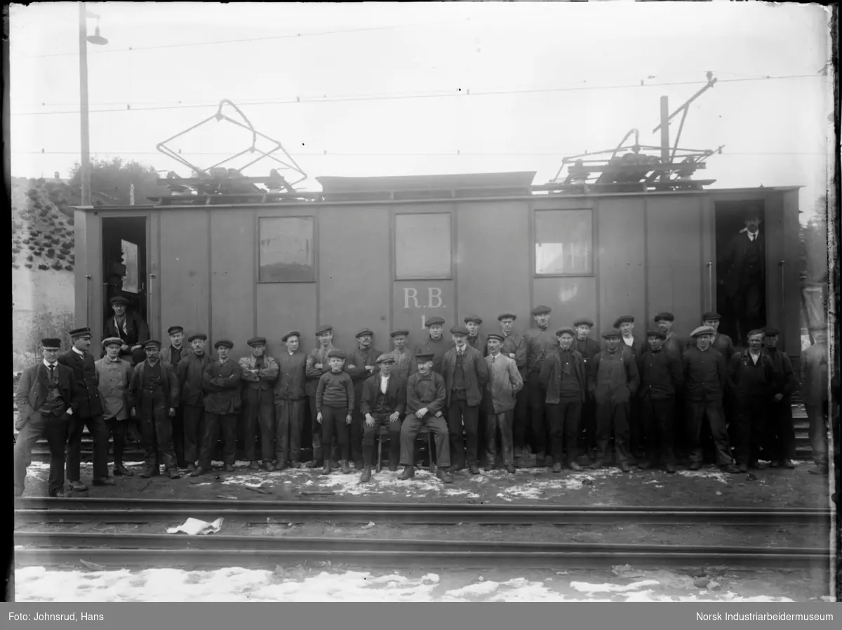En gruppe jernbanearbeidere fotografert foran et elektrisk lokmotiv ved Rjukanbanen Jernbaneverktsted. Lokomotivet er merket R.B. 1