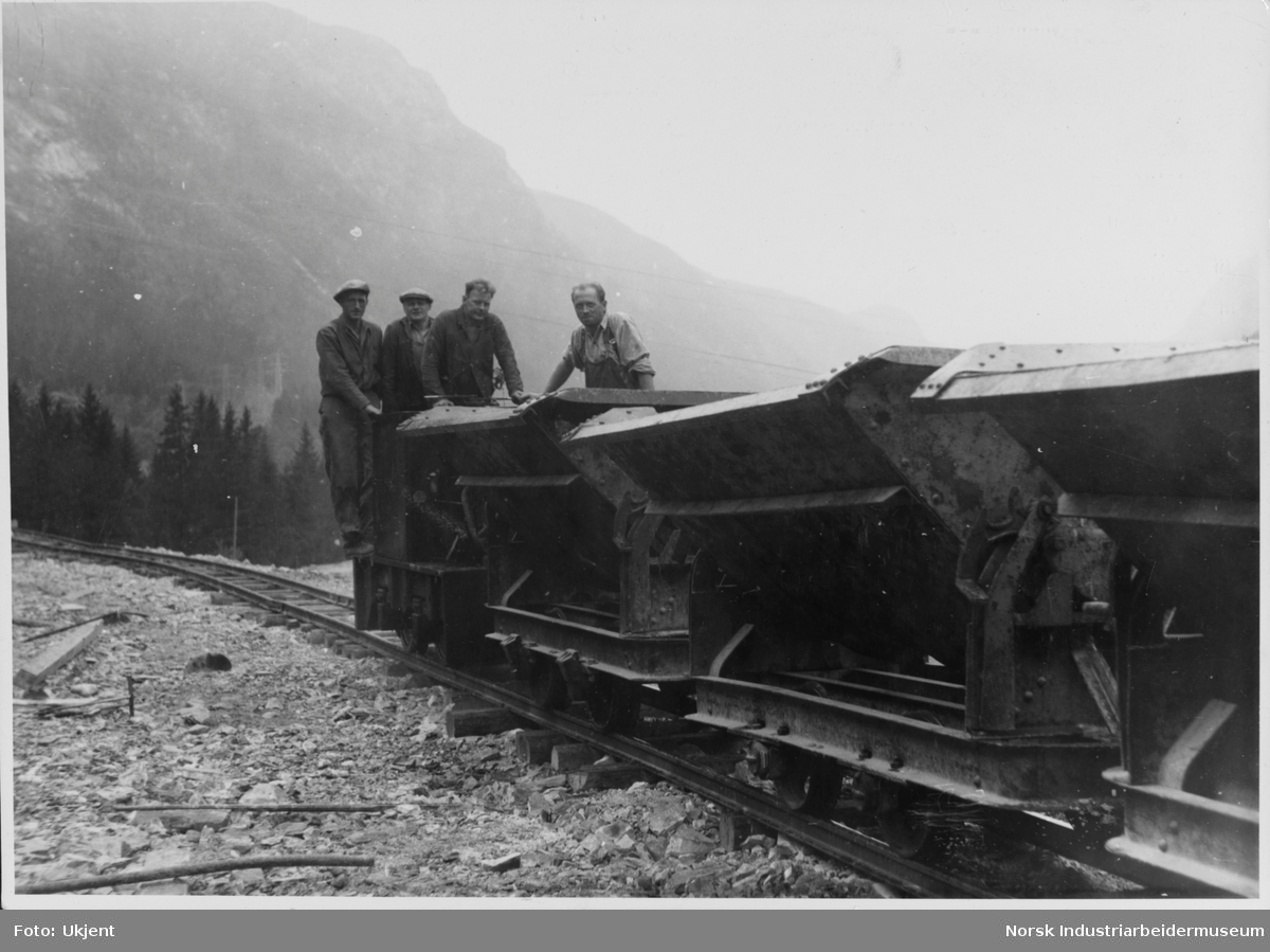 Anleggsarbeidere stående på jernbanevagger på jernbanespor, Måranlegget, under transport av stein ut av tunellen på tippen nederst i Svineroiveien.