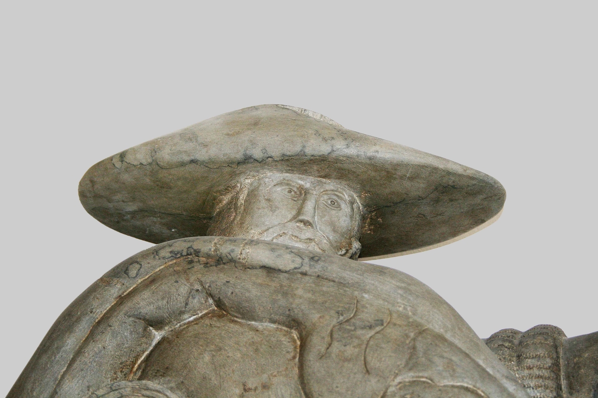 Väggskulptur i patinerad brons. Föreställer "Knallen". Gående, mustaschprydd man, i bredhättad hatt. På höger sida, bär han textilier och alnmått. I vänster hand, håller han en pingla. Proveniens Algot Johansson AB, Borås.

Funktion: Väggdekoration