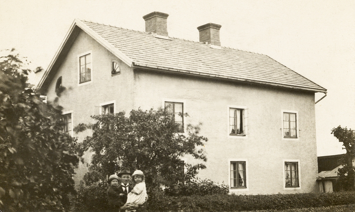 En okänd man står med två små barn i famnen framför ett bostadshus, ca 1920-25.