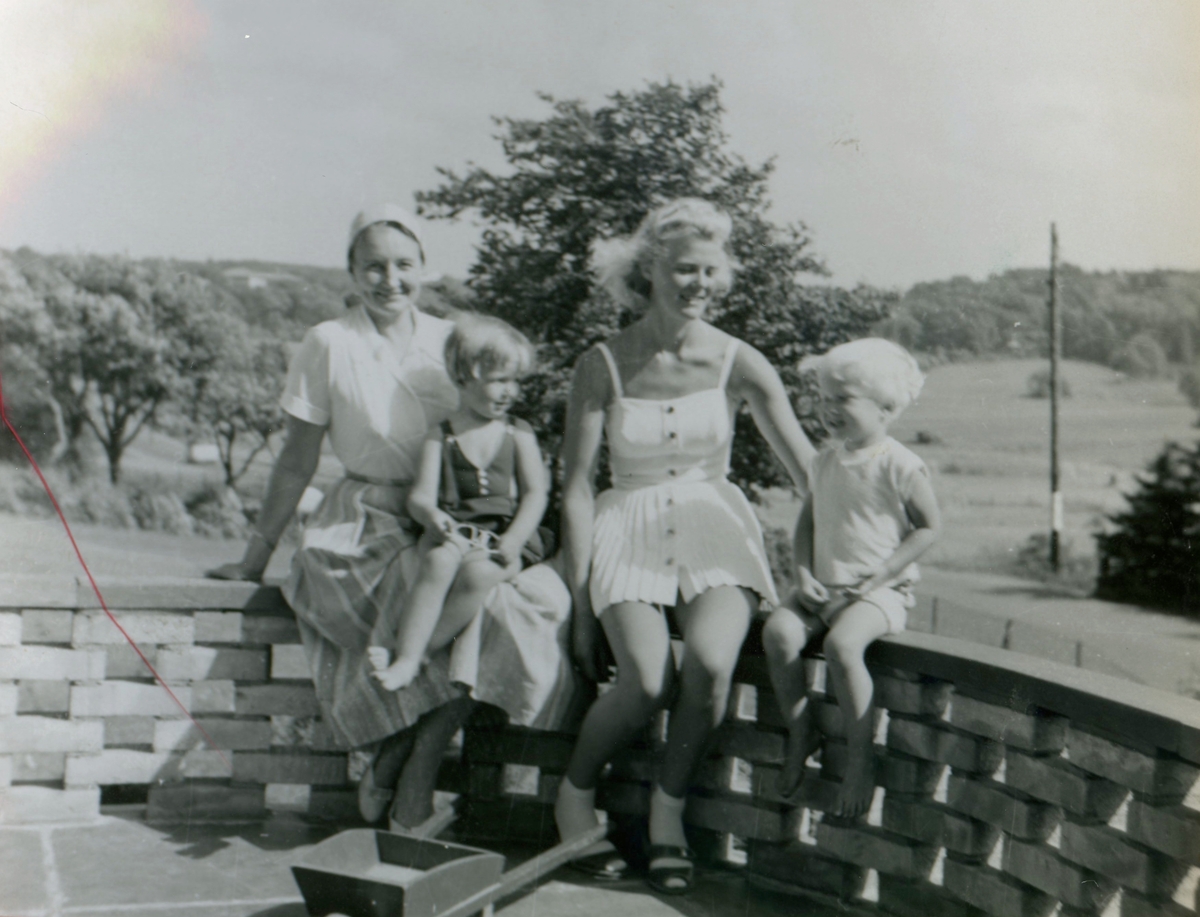 Två kvinnor och deras barn sitter utomhus på en altanmur, sommaren 1959 i Kållered. Från vänster: Mary Ekman (1920 - 1988) med sonen Manne (född 1955) i knät och grannen Lilian Sjöström (1932 - 1998) med sonen Sven-Göran (född 1955).