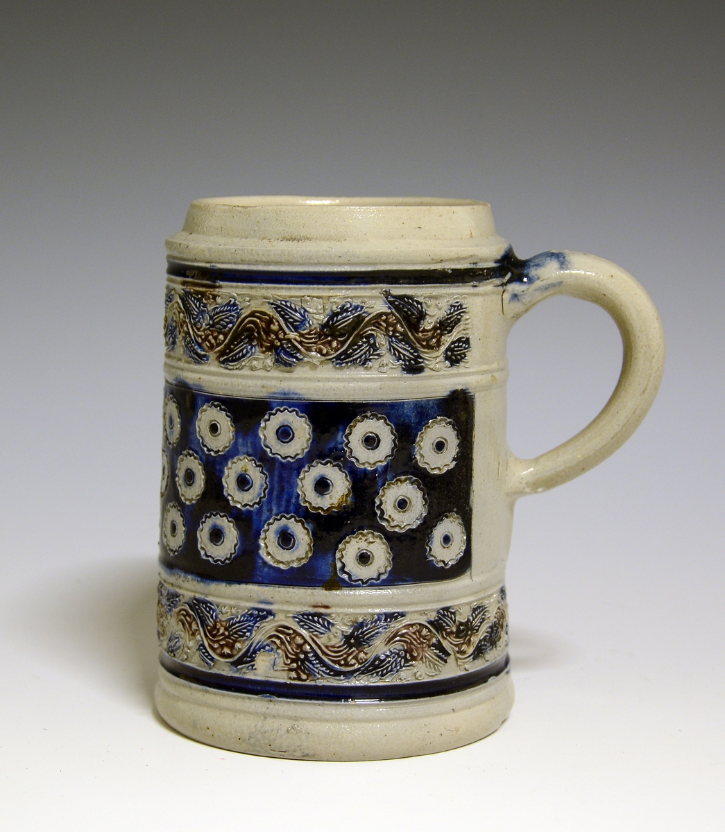 Ølkrus av steintøy med hank. Dekorert med border og mønster i relieff, i blått og manganrødt.

Prot: Tysk keramikk.
