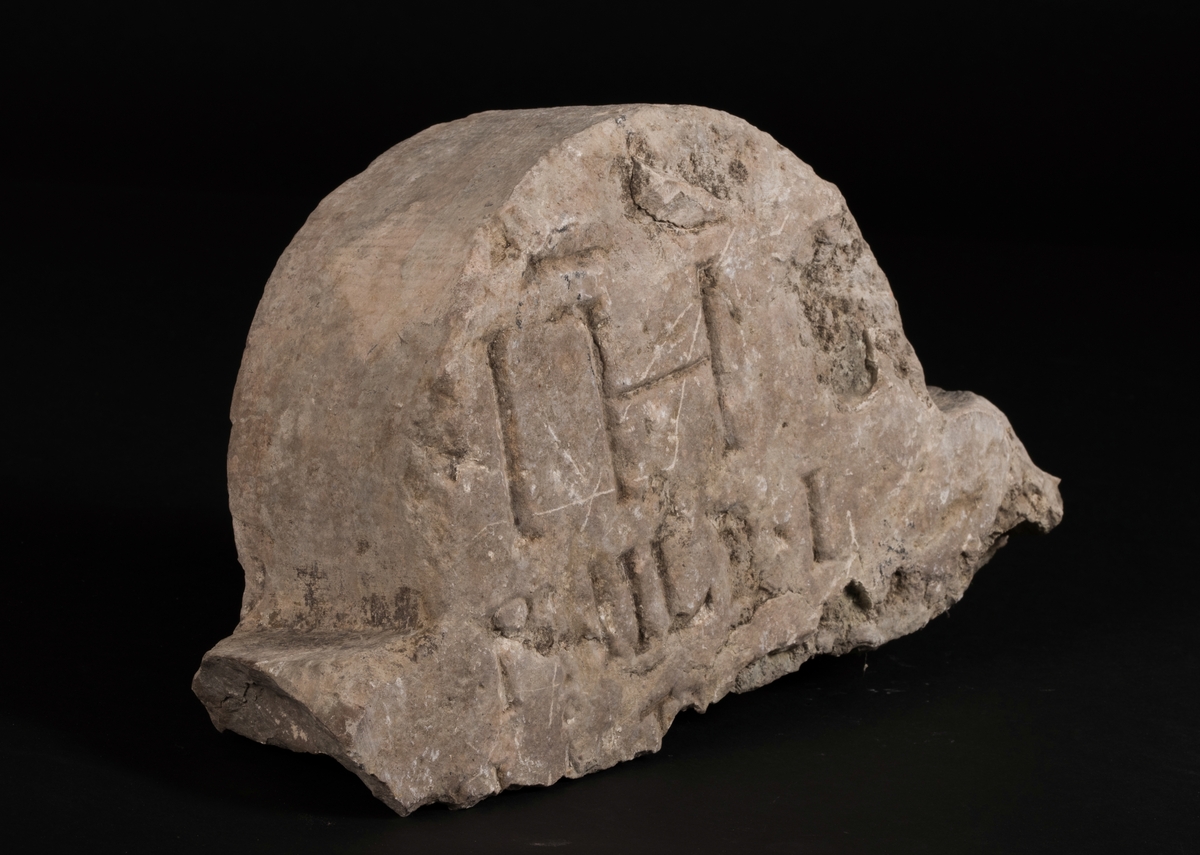 Gravstensfragment tillverkat av huggen kalksten.
Överdelen av en gravsten dekorerad med ett änglahuvud i relief. På baksidan inhugget "IH" samt troligen ett S och därunder fler bokstäver eller sifffror.