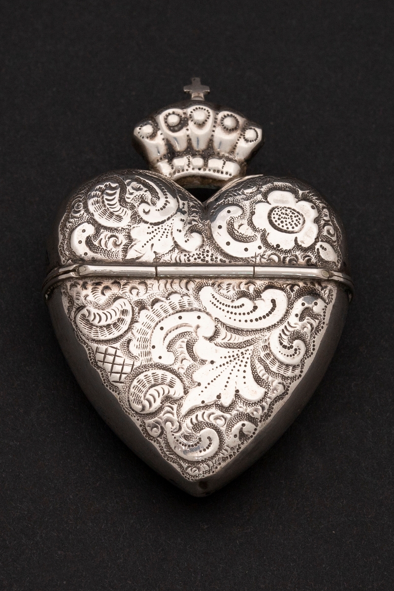 Hjerteformet luktevannshus i sølv med krone og kors på lokket. Rokokko-ornamentikk på for- og bakside av korpus. Kantsidene er uten dekor.