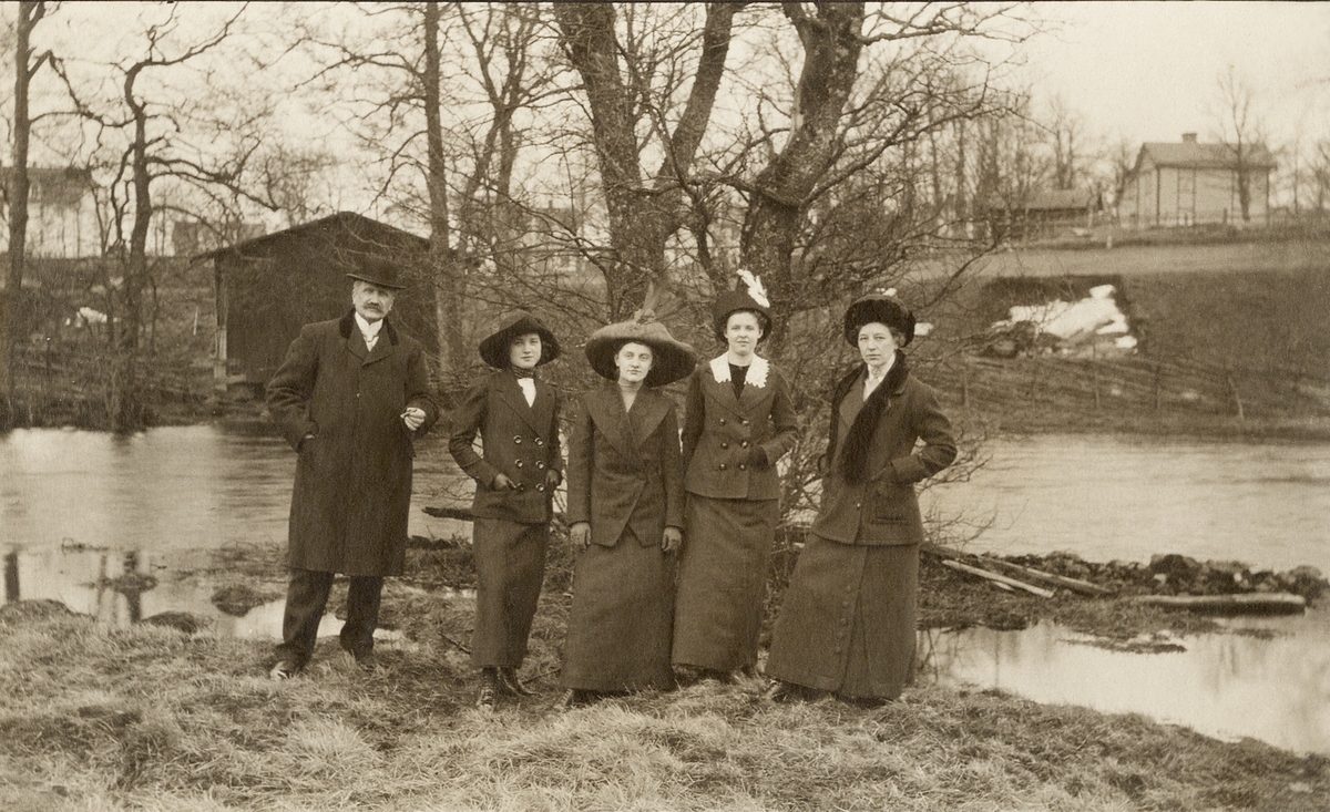 En medelålders man i paletå och hatt och en kvinna i promenaddräkt flankerar tre yngre kvinnor vid en åkant 
en vårvinterdag. Trol. i Åsedatrakten, ca 1915.