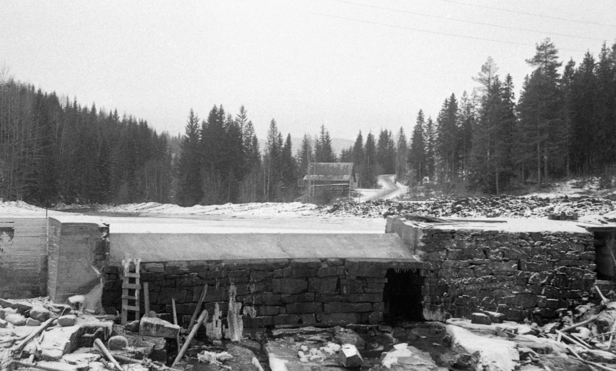 Mølledam, sannsynligvis ved Flaen mølle i Hurdalselva (Høverelva) i Akershus etter reparasjon i 1957. Fotografiet er antakelig tatt seinhøstes, etter at det hadde kommet litt snø og på et tidspunkt da vannføringa i elva var minimal. Vi ser dammen i motstrøms retning. Det dreide seg om en tørrsteinsmurt damkonstruksjon der overløpssona på midten ser ut til å ha blitt påstøpt en terskel av betong, antakelig for å øke volumet på det bakenforliggende vannreservoaret. Også damarmen til venstre framstår som en støpt konstruksjon, som kan ha vært ny da dette bildet ble tatt. Helt til høyre i bildet skimter vi en mindre åpning i dammen. Dette kan ha vært vanninntaket til den nevnte mølla. I sona mellom vassdraget og skogen til høyre i bildet skimter vi en veg. Ved vegen lå det en uthusbygning.