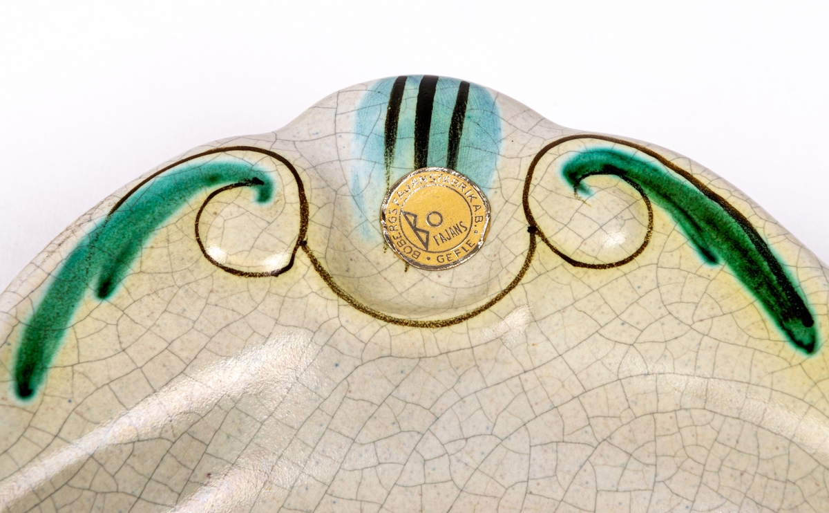 Fyrkantig assiett i lergods från Bo Fajans, modell 1084, formgiven av konstnär Allan Ebeling 1925. Glasyr KK: gråvit antik konstglasyr (krackelerad) med handmålad dekor i grönt, blått, brunt och gult, med krusbärsliknande bär i spegeln och blad- och streckdekor på brämen.