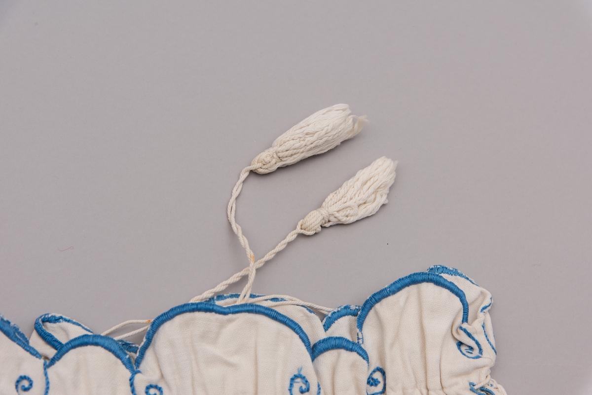 Hvit tekstilpose med blått broderi. Det er dusker på enden av tråden.