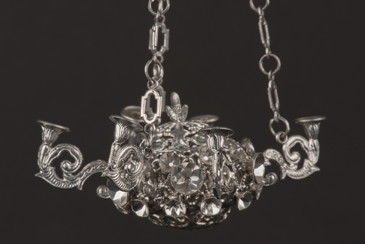 Ljuskrona till dockskåp, tillverkad av tenn.
Kronan har 6 ljusarmar, något tillböjda, och hänger i 3 kedjor.