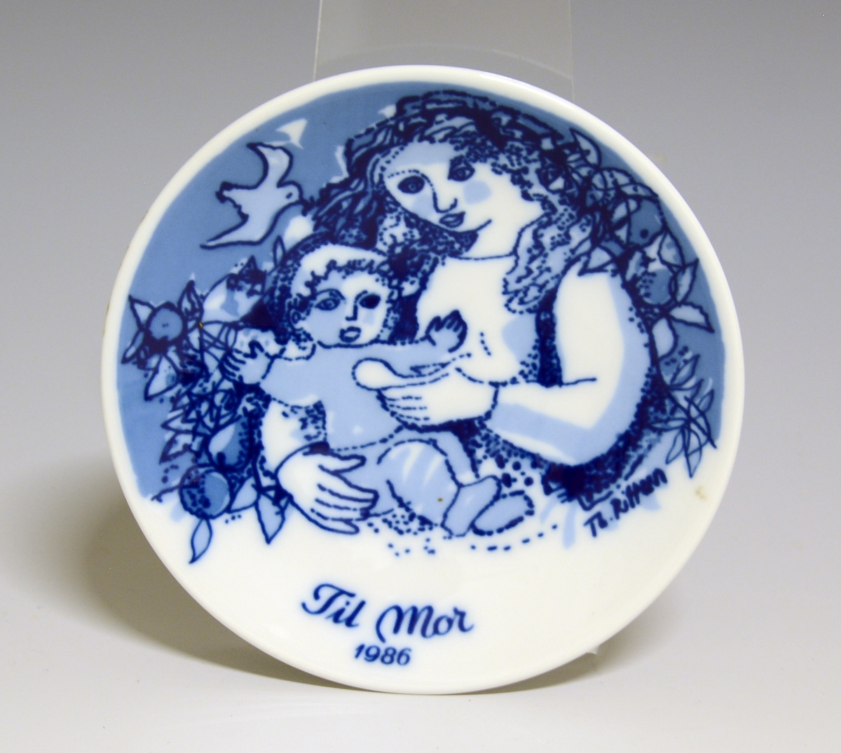 Morsdagsplatte av porselen. Hull til oppheng bak. Hvit glasur. Dekorert i blått med sittende kvinne som mater et lite barn, tekst Mors Dag 1986.